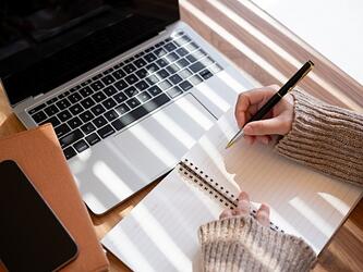 person's hands using notebook and pen alongside laptop on desk